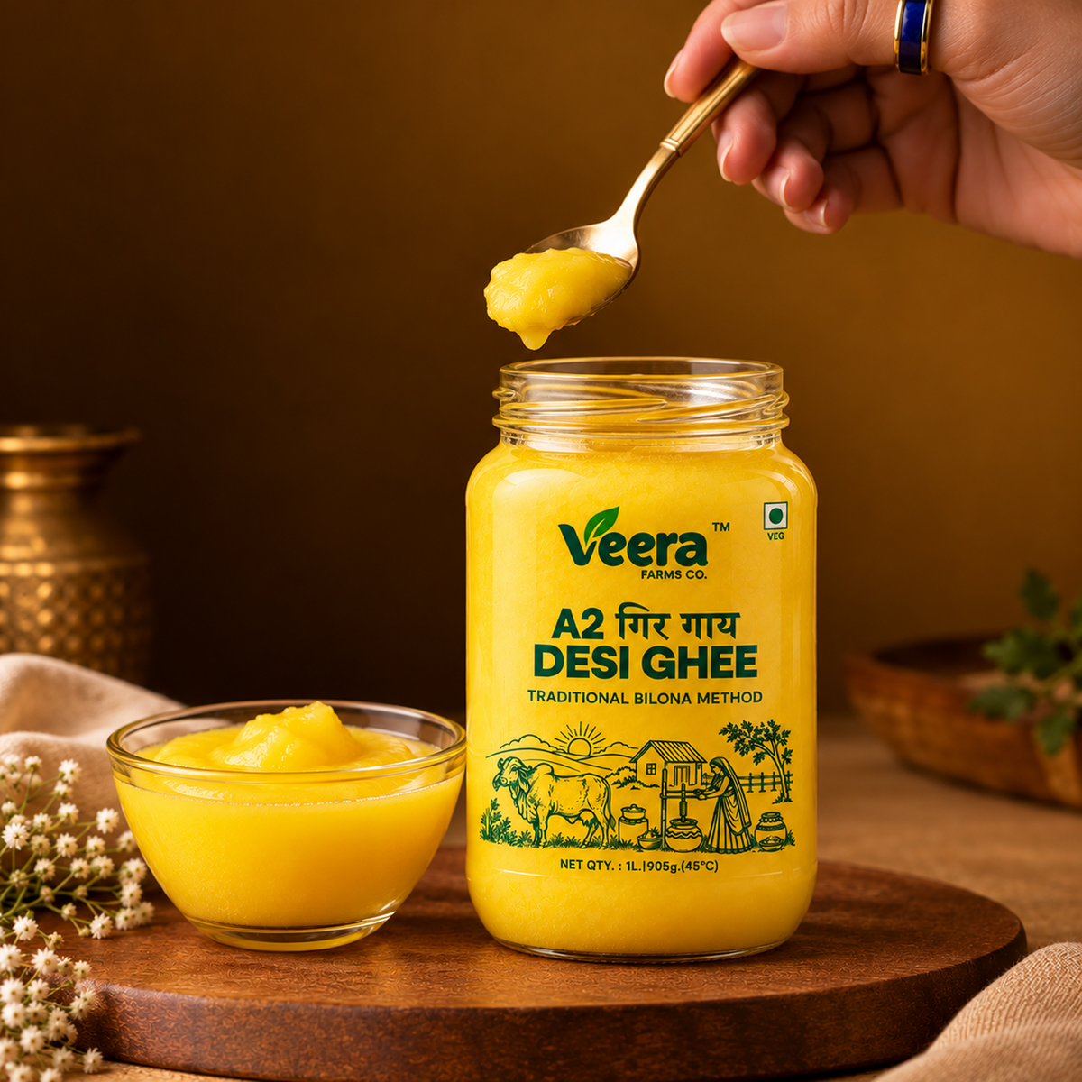 A glass jar of Veera Farms Co. A2 Desi Ghee sits on a wooden board, with a hand holding a spoonful of the golden yellow ghee above the open jar.
